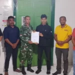 Six men pose indoors as a soldier in camouflage hands a document to a young man in black others watch the handover